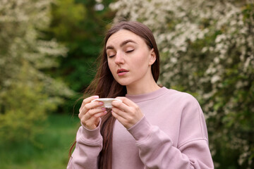 Woman with paper tissue suffering from seasonal pollen allergy near blossoming shrub on spring day
