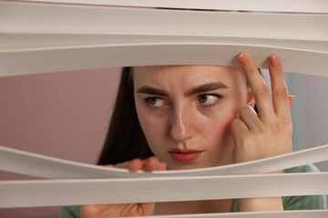 Paranoia. Worried woman looking through window blinds indoors