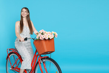Smiling woman posing on bicycle with basket of flowers against light blue background. Space for text