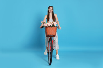Smiling woman riding bicycle with basket of flowers against light blue background