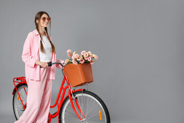 Smiling woman in sunglasses and bicycle with basket of flowers against grey background. Space for...