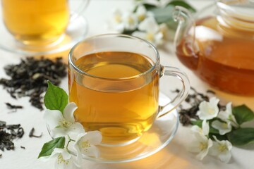 Aromatic jasmine tea in glass cup, teapot, flowers and dried leaves on white table, closeup