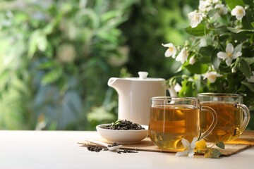 Tasty jasmine tea in cups, teapot, dry leaves and flowers at white wooden table, closeup. Space for text