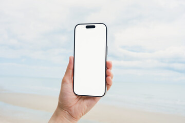 Hand holding a smartphone with a blank screen, on the sea and sandy beach background. Summer vacation concept.