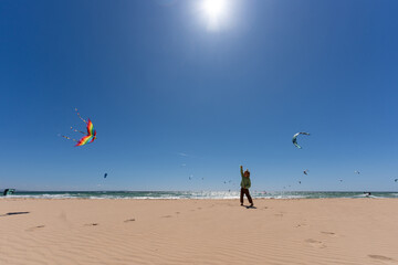 Obraz premium Young child flying a colorful rainbow kite on a sunny beach with a clear blue sky and kitesurfers in the background. Concept of summer fun, childhood joy, and outdoor freedom