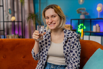 Satisfied blonde Caucasian woman sitting at home on sofa couch leading healthy lifestyle, drinking a glass of pure water, smiling. Drink daily norm of water. Girl smiling at camera in apartment home.