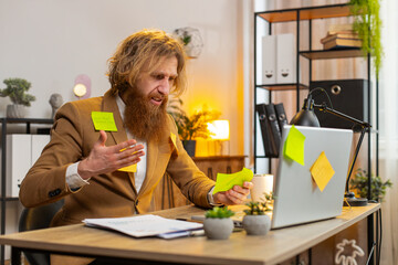 Tired exhausted frustrated bearded businessman sits in home office table with laptop sticky notes...