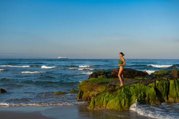 Slim woman in green swimsuit stands calmly on mossy rocks by the ocean at golden hour, gazing into...