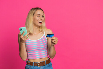 Happy Caucasian young blonde woman in crop top using credit bank card smartphone while transferring...