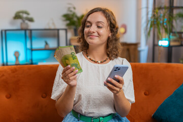 Successful rich woman holding smartphone and waving euro bill money fan sitting on sofa couch at...