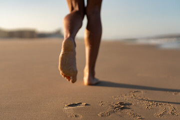 Close-up of toned female legs walking barefoot along the sandy beach in soft morning light, leaving footprints behind on the warm, wet sand with gentle ocean blur in background.