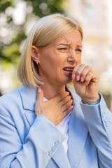 Sick mature businesswoman in formal suit suffering from sore throat while standing on city street....