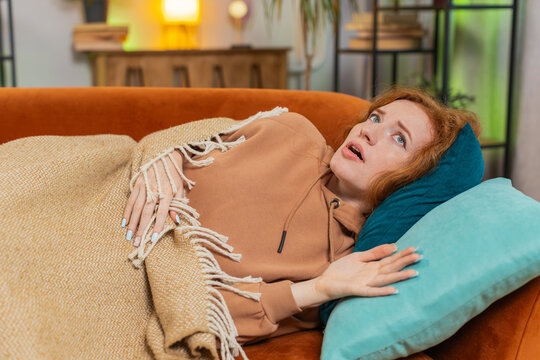 Young Caucasian woman suddenly wakes up from nightmare while sleeping on couch due to anxiety, disorder, bad dreams and illness or disease. Tired girl trying to sleep on sofa in living room at home.