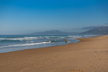 Woman walking along an empty beach with ocean waves and mountains in the background, peaceful coastal landscape with footprints on sand under clear blue sky, early morning light.