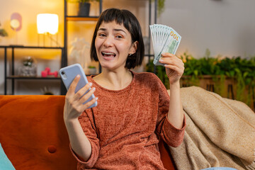 Smiling young woman counting money dollar banknotes using smartphone calculator on sofa in room at...