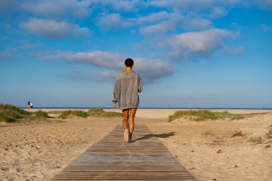 Woman in oversized sweater walking barefoot on a wooden path toward the beach, peaceful morning scene with cloudy blue sky and distant ocean horizon. - Powered by Adobe
