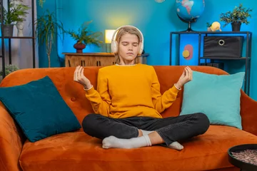  Caucasian teenager boy meditates in lotus pose on comfortable sofa at home. Wearing headphones listening music, child relaxes unwinds peacefully after long school day, finding inner peace calmness © Andrii Iemelianenko