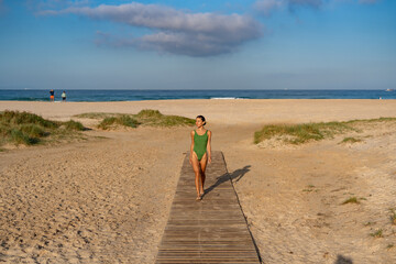 Confident woman walking barefoot along wooden beach walkway in green swimsuit, surrounded by golden...