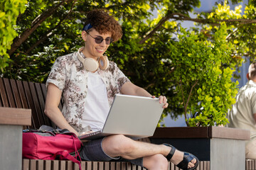 Happy young man working online distant job with laptop in city sunshine street sitting on bench,...