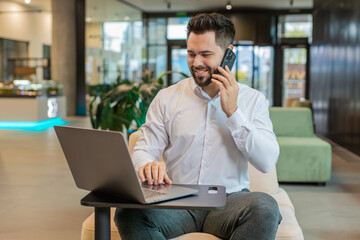 Fototapeta na wymiar Young businessman in formal shirt answering to client on mobile phone call, using smartphone and laptop. Conversation. Caucasian bearded man guy working online job, talking in modern office lobby.