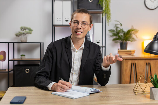 POV shot of young businessman freelancer looking to camera listening to colleague talking writing down on diary in video chat at home office. Stylish guy in suit taking notes at table. Web cam view.