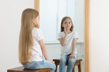 Upset little girl looking at herself in mirror indoors