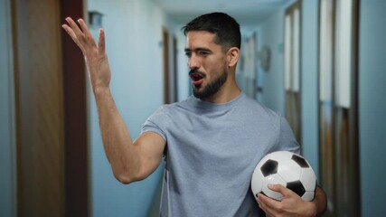 Hispanic man in grey shirt holding soccer ball in hotel hallway with wooden doors and blue walls, expressing various emotions and gestures, showcasing diverse mood dynamics. - Powered by Adobe