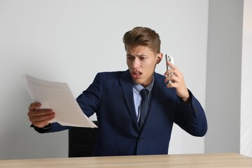 Young businessman with papers complaining to someone via phone at wooden table indoors
