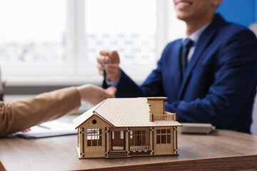 Real estate agent giving key to client at desk in office, focus on house model