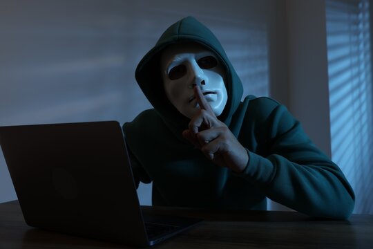 Anonymity. Masked man with laptop showing hush gesture at table in dark