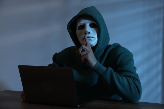 Anonymity. Masked man with laptop showing hush gesture at table in dark