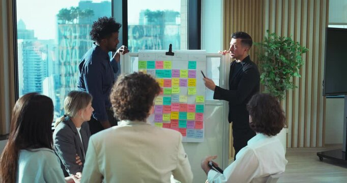 Diverse group of professionals discussing ideas with sticky notes on a whiteboard in a bright, modern office meeting room.