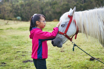 Young girl wearing glasses and a pink jacket gently caresses a white horse in a vibrant green meadow