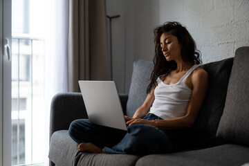Freelance woman working from home on laptop, sitting comfortably cross-legged on a couch near...
