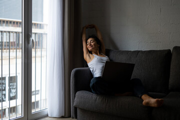 Young freelance woman in casual clothes smiling and stretching with hands behind head, relaxing on gray sofa after finishing remote work on laptop at home.