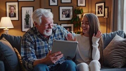 happy granddaughter and grandfather are having fun to make a selfie or video technology call to family and relatives with tablet while sitting on sofa in living room at home - Powered by Adobe