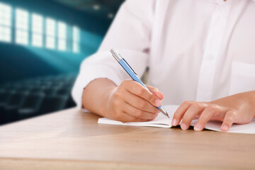 Close up of boy hand writing in notebook at school desk with blurred classroom or university hall...