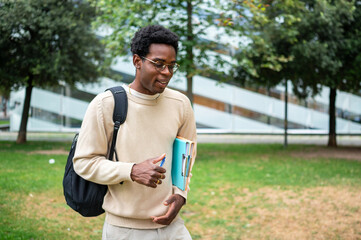 Smiling university student walking in campus park holding folders and pen