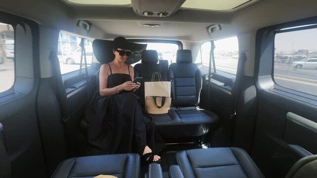 Fashionable woman in a black dress and hat is seated in a luxurious vehicle, checking her phone