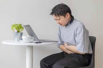 Asian man suffering from a stomach ache or period cramps while working on a laptop. Concept of...