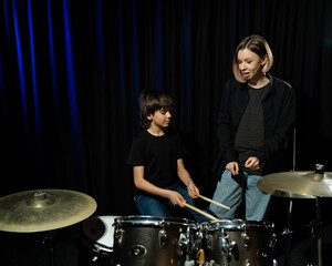 Young caucasian woman teaches a boy to play the drums in the studio on a black background. Music...