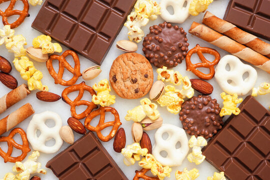 Background of various sweet and salty snacks including chocolate, popcorn, pretzels, nuts and cookies. Above view over a white background.