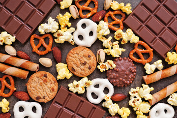 Background of various sweet and salty snacks including chocolate, popcorn, pretzels, nuts and cookies. Top down view over a dark background.