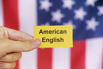 A young student holding a yellow piece of paper with the words American English on it in front of the American flag.