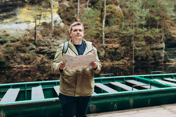 Tourist man studies map while on wooden pier by river. Traveler with backpack, planning outdoor...