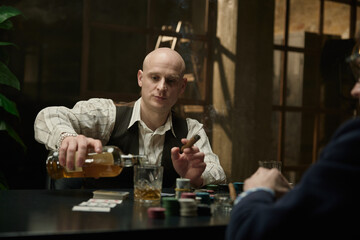In a dimly lit room, bald man confidently pours whiskey while participating in poker game