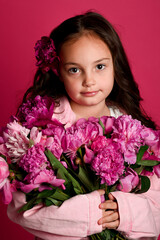 A young girl of Caucasian descent holds a bouquet of vibrant pink peonies against a bright pink...