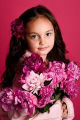 A young girl of Caucasian descent smiles, holding a bouquet of vibrant pink flowers against a...