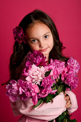 A young girl of Caucasian descent holding a bouquet of vibrant pink peonies against a bright pink...