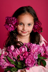 A young girl of Hispanic descent holds a vibrant bouquet of pink peonies against a bright pink...